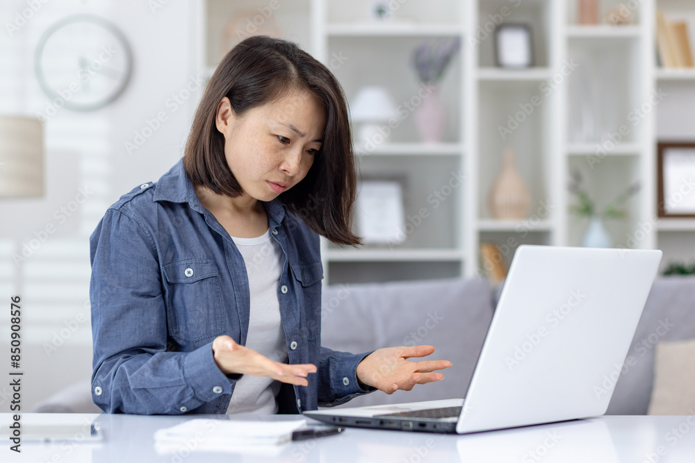Frustrated and serious young Asian woman sitting in the office in casual clothes, working on a laptop, looking disappointed at the screen and spreading her hands.