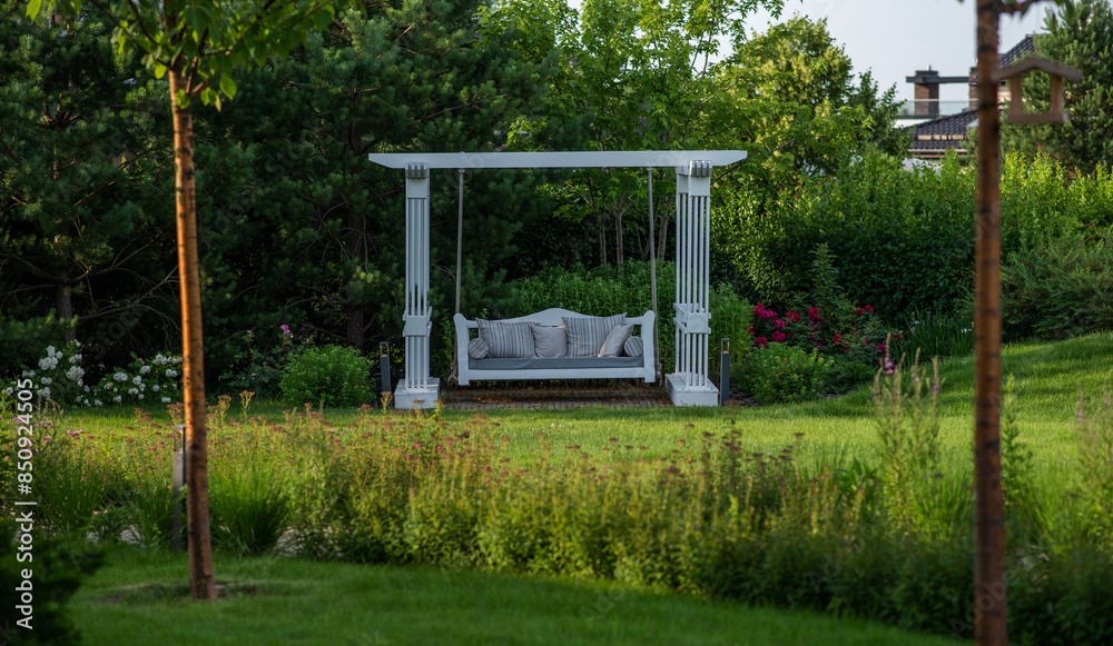 Fototapeta premium A tranquil white garden swing bed nestled among lush greenery in a peaceful garden, offering a picturesque spot for relaxation amid blooming roses and verdant shrubs