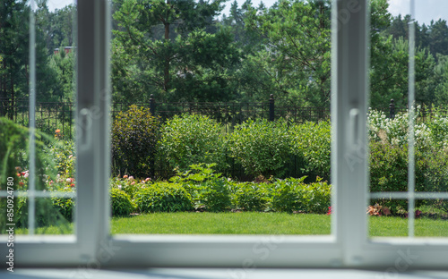 Through the panes of a window, reveals a meticulously cared for garden, resplendent with a variety of shrubs and blooming flowers, and flanked by tall trees, offering a slice of nature's tranquility.