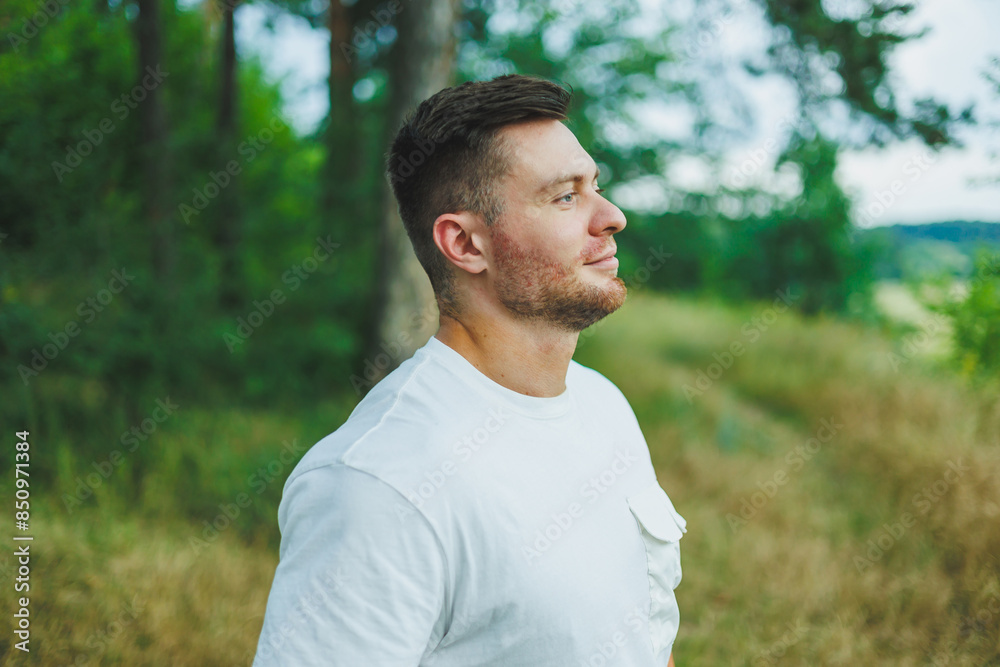 A man in a white T-shirt with a laptop stands in nature and looks into the distance. A man rests in nature from work.