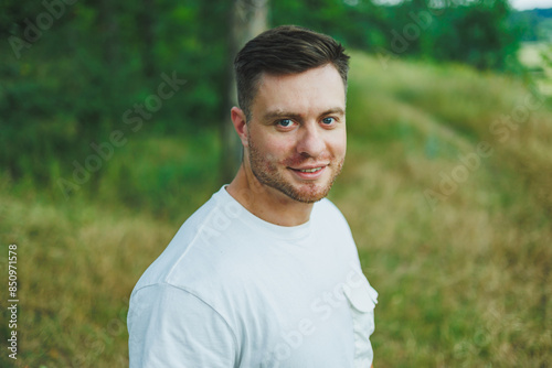 A man in a white T-shirt with a laptop stands in nature and looks into the distance. A man rests in nature from work.