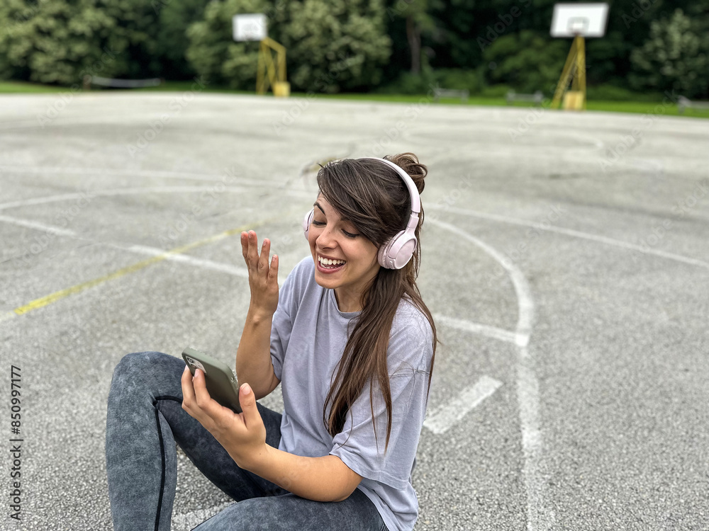 Fototapeta premium Young woman wearing headphones using mobile phone for video call, gesturing hi to friends or parents after exercise, jogging run in city park. counting calories