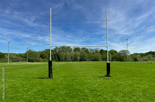 A pair of rugby goal posts stand prominently at the center of a lush, green field under a blue sky. In the background, trees line the edge of the playing area in, Cottingley, UK