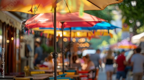 A row of outdoor cafe tables with colorful umbrellas overhead. People are enjoying drinks and conversation in the background. The scene is bustling with activity.