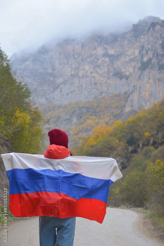 Woman traveler on a winding road in the mountains on a foggy autumn day with the flag of the Russian Federation on her shoulders