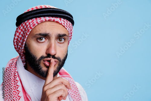 Man dressed in muslim clothes asking to keep secret while holding finger on lips and looking at camera. Arab person wearing headscarf with rope band showing silent gesture portrait photo