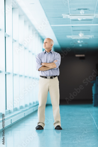Senior businessman standing with folded arms in office corridor