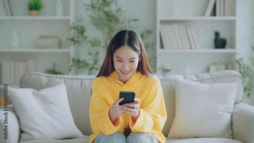 Happy young Asian woman relax sitting on couch using cell phone, smiling lady laughing holding smartphone, looking at cellphone enjoying doing online ecommerce shopping in mobile apps Happy young Asian woman relax sitting on couch using cell phone, smiling lady laughing holding smartphone, looking at cellphone enjoying doing online ecommerce shopping in mobile apps