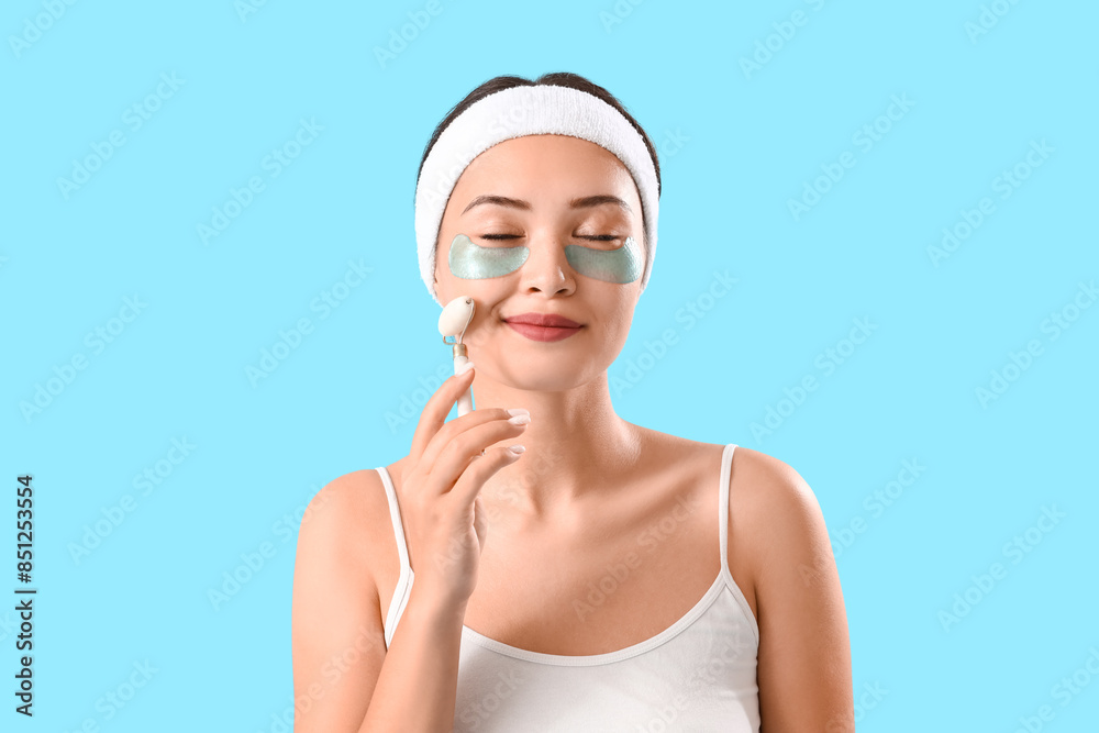 Pretty young woman with under-eye patches and facial massage roller on blue background Pretty young woman with under-eye patches and facial massage roller on blue background