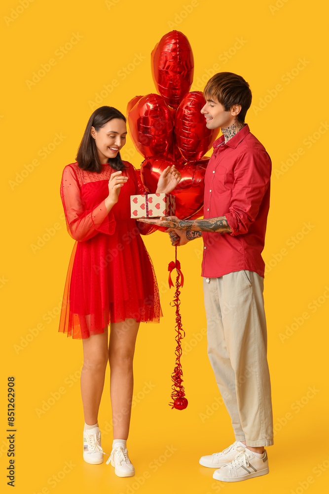 Young couple with air balloons in shape of heart and gift box on yellow background. Valentine's Day celebration Young couple with air balloons in shape of heart and gift box on yellow background. Valentine's Day celebration