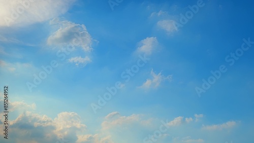 A bright blue sky with a few scattered white clouds. Some clouds have a slight golden tint from the sun, creating a warm and peaceful atmosphere. Blue sky background.
