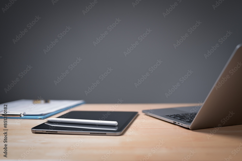 A laptop and tablet sit on a desk with a clipboard and pen. The desk is cluttered with papers and the laptop is on a wooden surface A laptop and tablet sit on a desk with a clipboard and pen. The desk is cluttered with papers and the laptop is on a wooden surface
