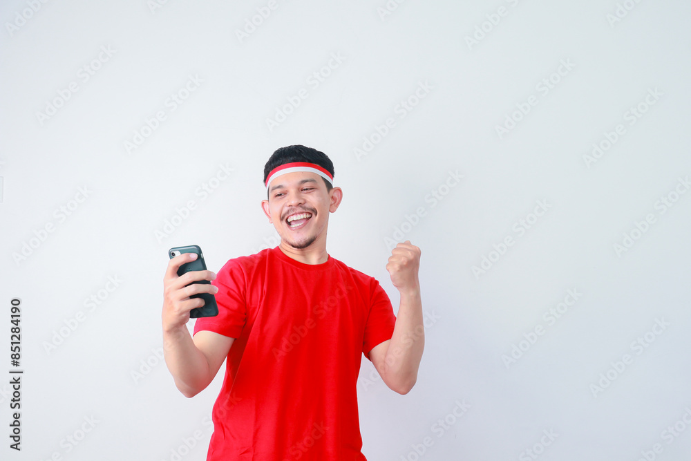 Excited young Asian man in casual t-shirt using mobile phone, getting good news isolated on grey background. indonesian independence day celebration concept Excited young Asian man in casual t-shirt using mobile phone, getting good news isolated on grey background. indonesian independence day celebration concept