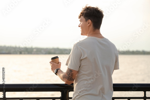 Handsome young man with cup of delicious latte by the waterfront, back view