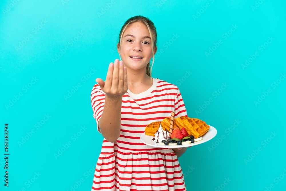 Little caucasian girl holding waffles isolated on blue background inviting to come with hand. Happy that you came Little caucasian girl holding waffles isolated on blue background inviting to come with hand. Happy that you came