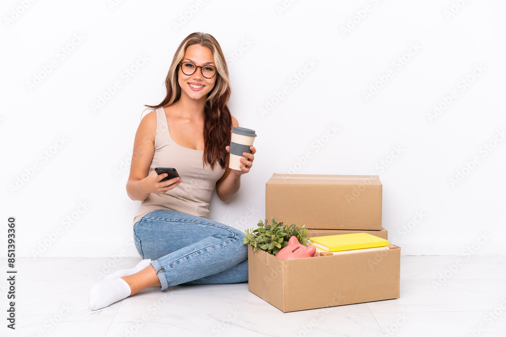 Young Russian girl sitting on the floor isolated on white background holding coffee to take away and a mobile Young Russian girl sitting on the floor isolated on white background holding coffee to take away and a mobile