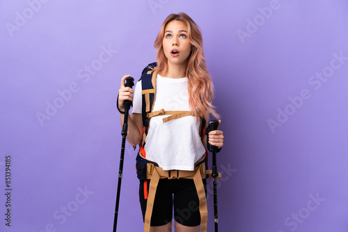 Teenager girl with backpack and trekking poles over isolated purple background looking up and with surprised expression
