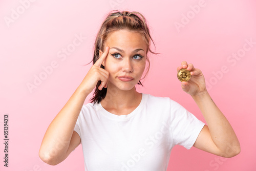 Teenager Russian girl holding a Bitcoin isolated on pink background having doubts and thinking