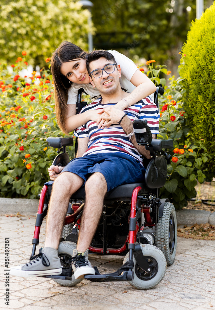 A man between the ages of 30 and 40 with a disability sits in an automatic wheelchair outdoors with a female caregiver.The woman hugs the disabled man from behind.Help for people with disabilities. A man between the ages of 30 and 40 with a disability sits in an automatic wheelchair outdoors with a female caregiver.The woman hugs the disabled man from behind.Help for people with disabilities.