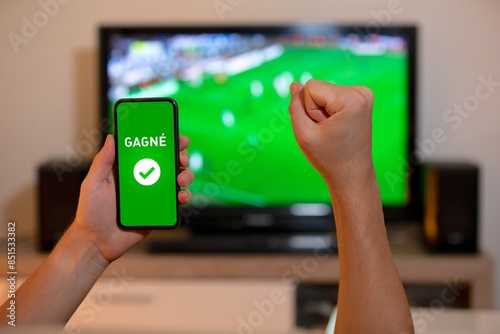 Un bras levé en signe de victoire pour un pari devant une télévision montrant un match de football, avec un téléphone affichant 