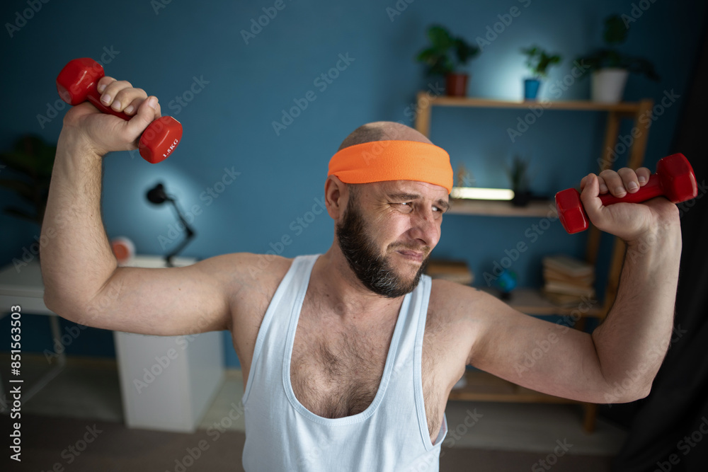 Middle-aged man lifting dumbbells at home, struggling with exercise, wearing orange headband and white tank top, blue background Middle-aged man lifting dumbbells at home, struggling with exercise, wearing orange headband and white tank top, blue background