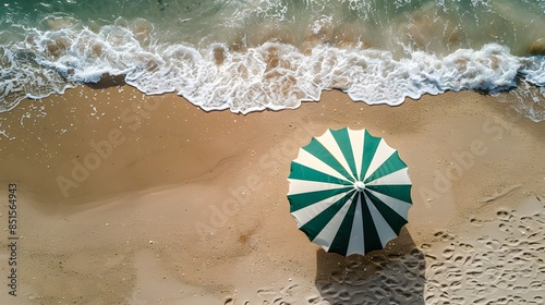 Aerial view of a green and white beach umbrella on a sandy shore with waves gently crashing. Ideal for summer and vacation concepts.