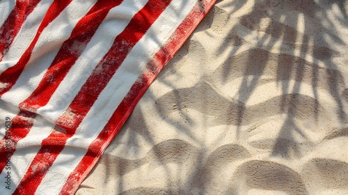Red and white striped towel on sandy beach, shaded by palm leaves, capturing the essence of summer relaxation and vacation vibes.