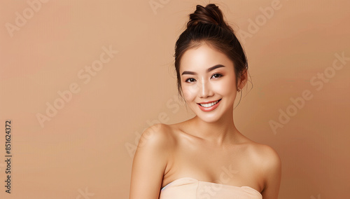A beautiful smiling Asian woman with perfect skin, in beige strapless dress and hair bun posing on light brown background for beauty portrait photography