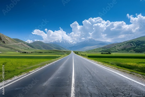 A wide road leads into the distance, with green fields on both sides and snow-capped mountains in the background. The blue sky is clear, and white clouds float across it