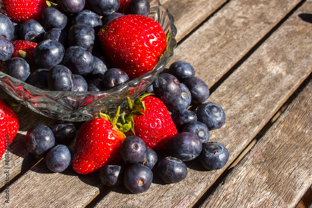A glass bowl, filled to overflowing with bright red strawberries and indigo-colored blueberries on a weathered wooden surface A glass bowl, filled to overflowing with bright red strawberries and indigo-colored blueberries on a weathered wooden surface