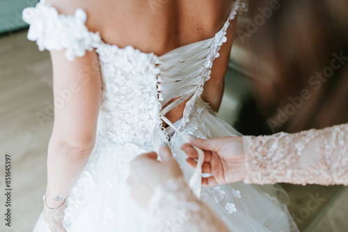 A bride or bridesmaid carefully adjusts the laces or buttons of a wedding dress, highlighting the detailed preparations and anticipation before the ceremony. photo