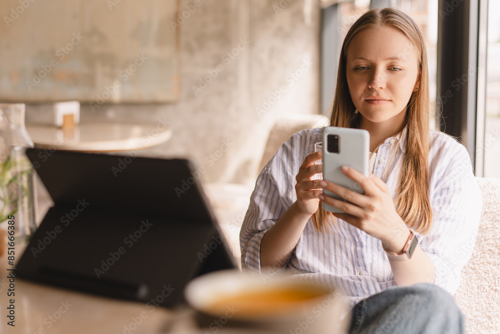 Young beautiful woman holding mobile and looking at smartphone while sitting at cafeteria. Calm girl using mobile phone and look flirting. Woman drinking water and smiling, sitting in cafe. Young beautiful woman holding mobile and looking at smartphone while sitting at cafeteria. Calm girl using mobile phone and look flirting. Woman drinking water and smiling, sitting in cafe.