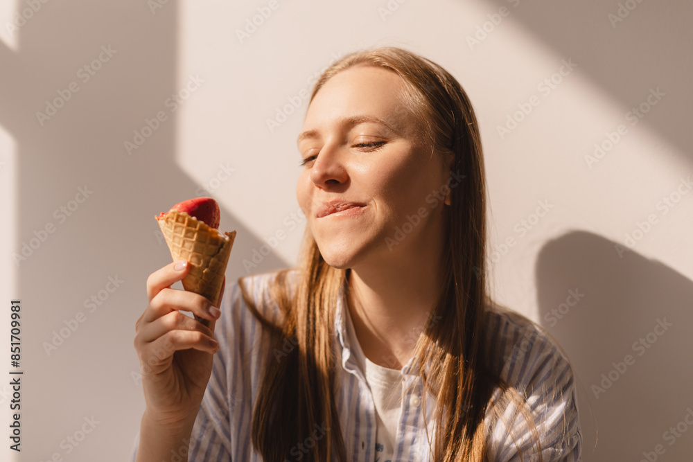 Close up portrait blonde woman hold delicious pink ice cream in waffle cone isolated white wall with sun light. Girl wear shirt in sunny day. Female lick ice cream and laughing, bite lips. Close up portrait blonde woman hold delicious pink ice cream in waffle cone isolated white wall with sun light. Girl wear shirt in sunny day. Female lick ice cream and laughing, bite lips.
