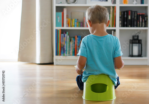 Konstfotografi Rear view of a 3-year-old Caucasian boy in light blue t-shirt sitting on green potty reading book, large bookcase full of books in background