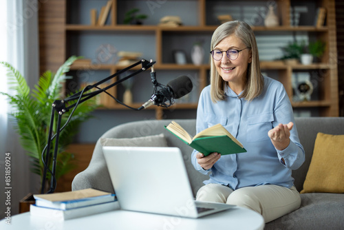 Senior woman recording podcast at home, holding a book and smiling, seated in front of a laptop and microphone.