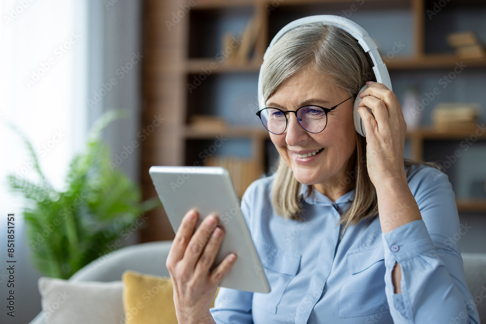 © Liubomir - Happy senior woman wearing headphones, using a digital tablet while sitting at home. Enjoying technology and media in a comfortable, modern environment.