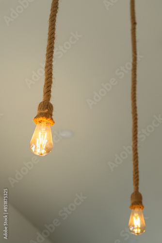 Rope ceiling lighting bulb glowing in orange warmlight shade, which is installed at living room for cozy design. Interior decoration object photo, close-up and selective focus. photo