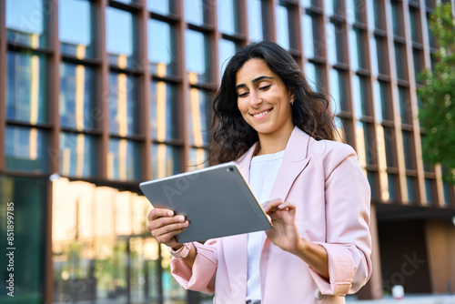 Smiling young middle eastern Israel businesswoman using tablet pc application for online remote work at office business building outdoors. Indian or arabic woman holding digital computer. Copy space