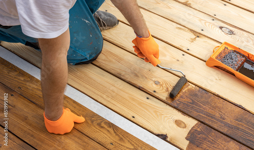 Wallpaper Mural A person wearing gloves uses a paint roller to apply wood stain to a wooden deck. The deck is made of light-colored wood and has a white border. Torontodigital.ca