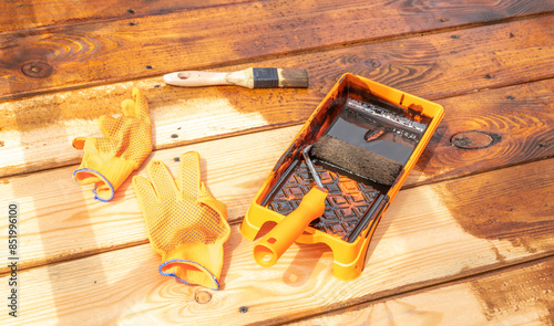 A close-up shot of a paint tray, gloves, and brush resting on a wooden deck, showcasing the process of staining the wood.