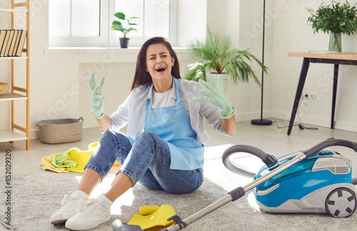 Young upset woman sitting on the floor in the living room at home next to vacuum cleaner tired of doing housework. Screaming girl vacuuming carpet in house frustrated with much housework
