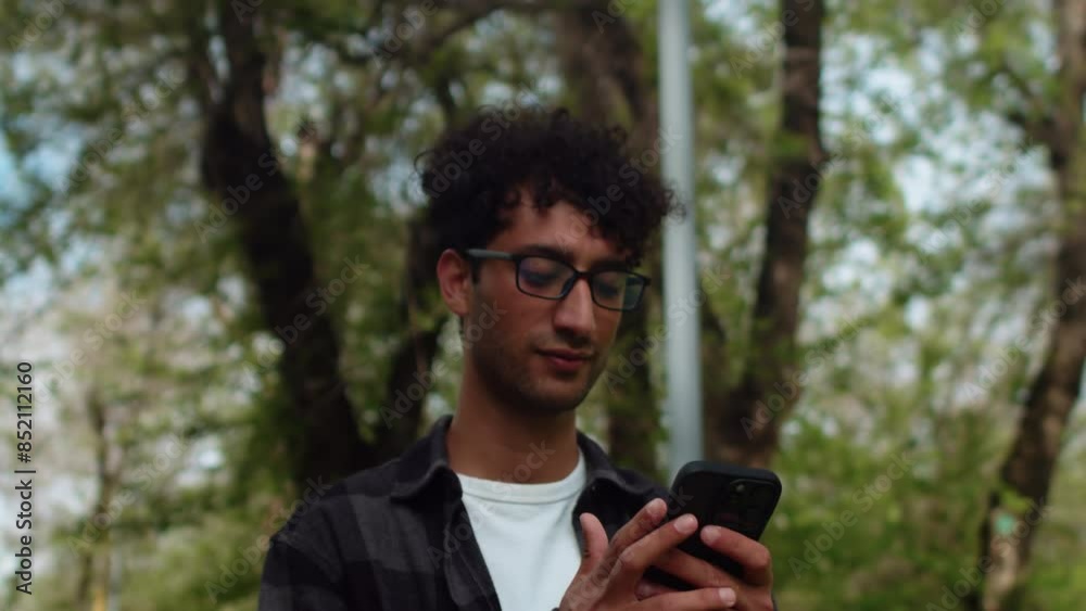 Young curly man in glasses and casual clothes close up walking in an alley of the city park, looking at his smartphone and smiling. Concept of carefree urban life on a sunny day Young curly man in glasses and casual clothes close up walking in an alley of the city park, looking at his smartphone and smiling. Concept of carefree urban life on a sunny day