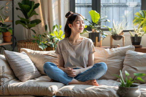 Calm Young Woman Meditating at Home Surrounded by Lush Green Plants While Sitting on a Comfortable Sofa in a Bright and Relaxing Living Room for Serenity and Mindfulness Practice