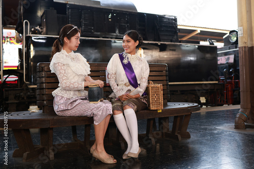 Two attractive women in traditional Thai dress pose for a photo with a steam train at the platform of the retro train station