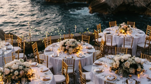 A sophisticated wedding setup on a seaside cliff, with round tables covered in white linens and gold chairs.  photo