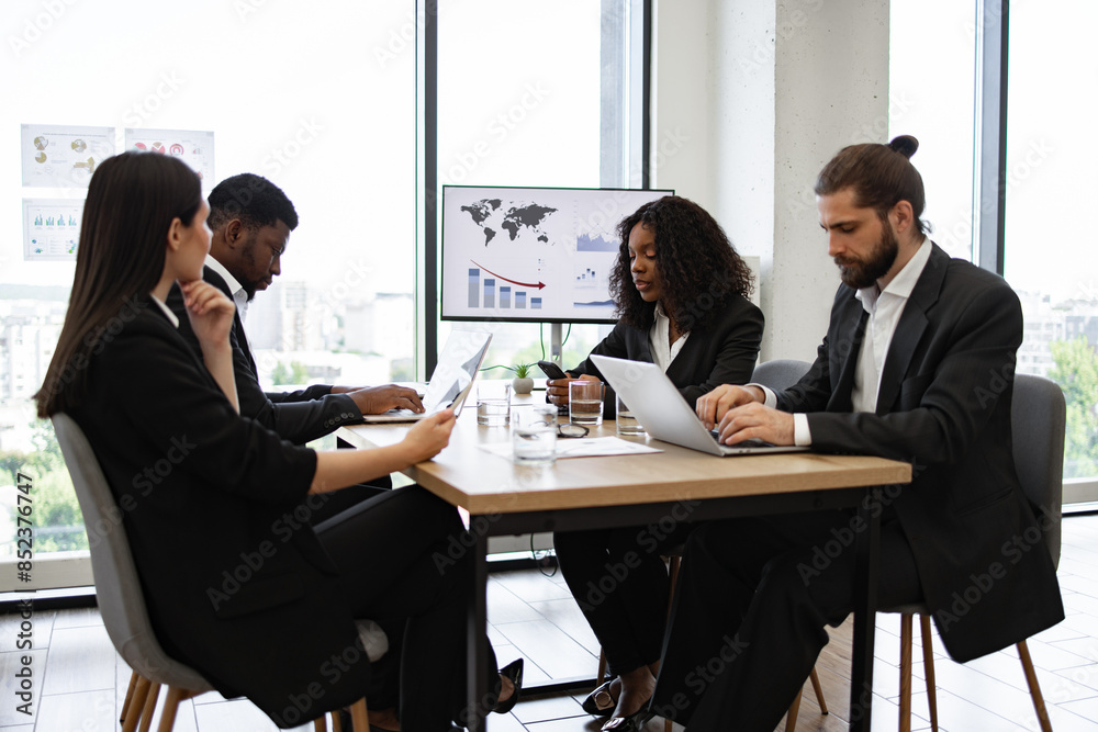Obraz premium Diverse group of business professionals engaged in analyzing data and collaborating in modern well-lit office. Team using laptops and digital devices for work with charts and graphs on screen.