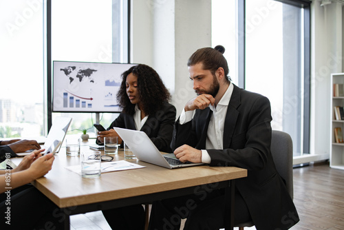 Group of professionals engaged in serious business meeting, using laptops, tablets, and smartphones, with presentation screen displaying charts in background.
