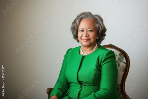 A smiling elderly Black woman in a green suit sits in a chair against a white background