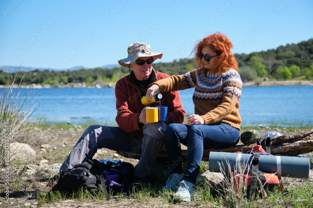 A man and a woman are sitting on a bench by a lake, enjoying a cup of coffee after hiking