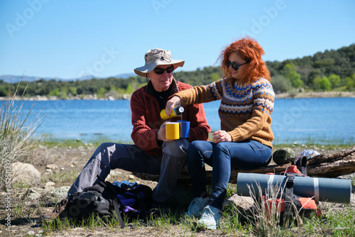A man and a woman are sitting on a bench by a lake, enjoying a cup of coffee after hiking